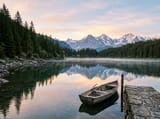 Photo of a calm alpine lake at sunrise with a pine forest, snowy peaks and a small rowboat at a stone jetty