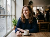 Photo of a young woman with wavy auburn hair and freckles in a navy knit sweater, seated at a wooden cafe table beside a window