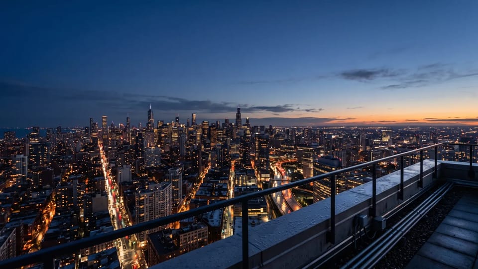 City skyline at blue hour seen from a rooftop railing, with lit buildings, glowing streets below, and an orange glow on the horizon